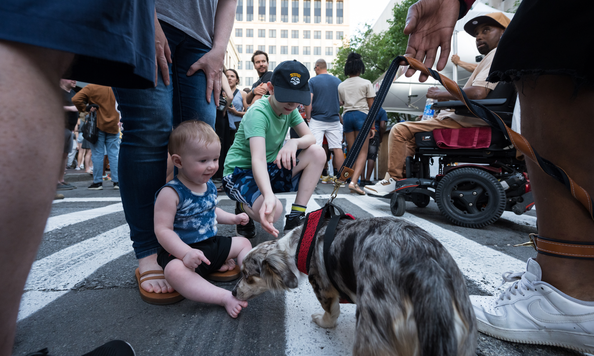 A baby and a small child pet a dog outside in the middle of a busy crowd