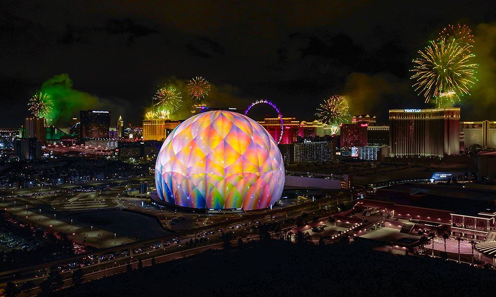 Night-time view of large multicolored sphere in center of Las Vegas downtown with fireworks in background.