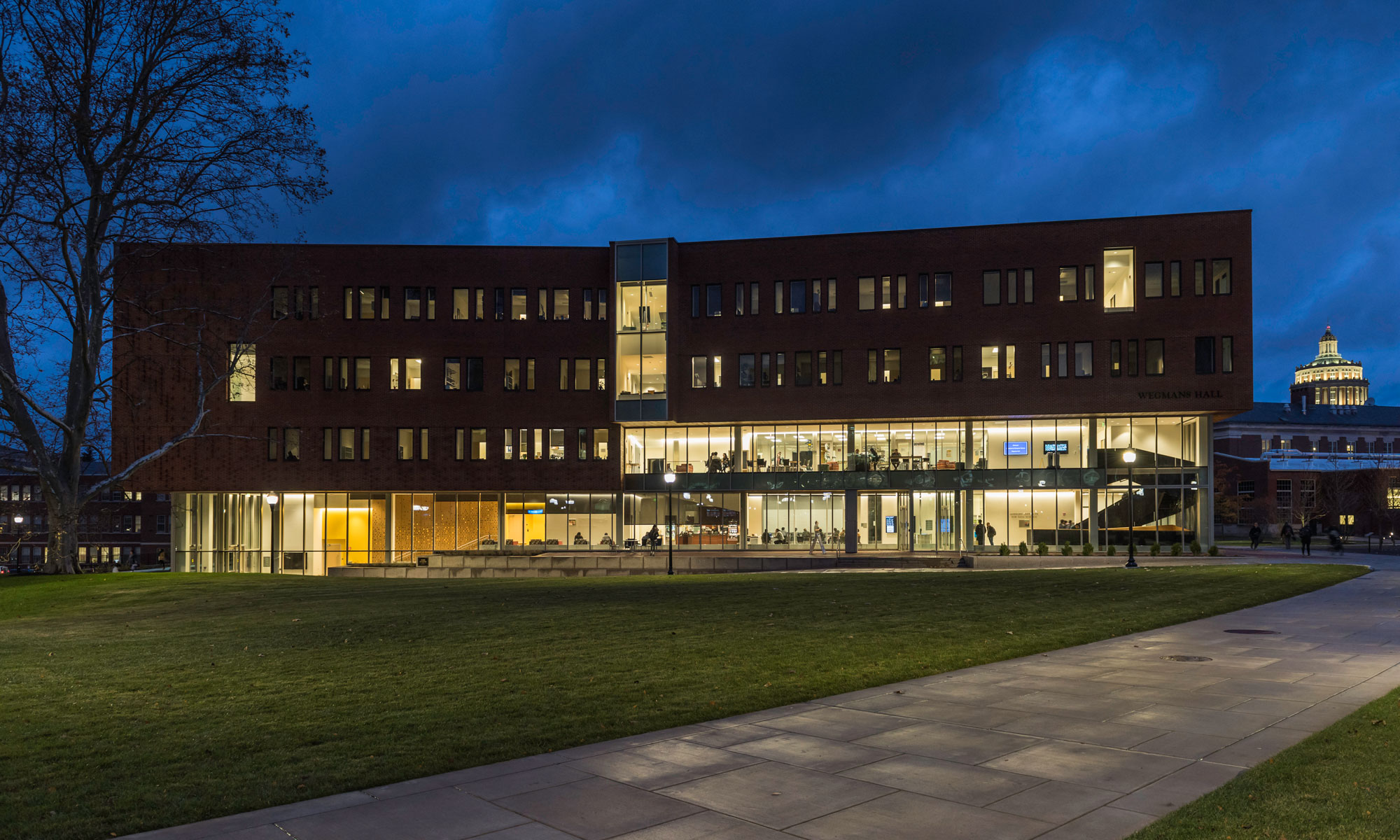 Wegmans Hall, a modern four-story brick building with large glass windows, lit from inside, at dusk. A well-maintained lawn with a walkway is in the foreground, and the University of Rochester Library is visible in the background.