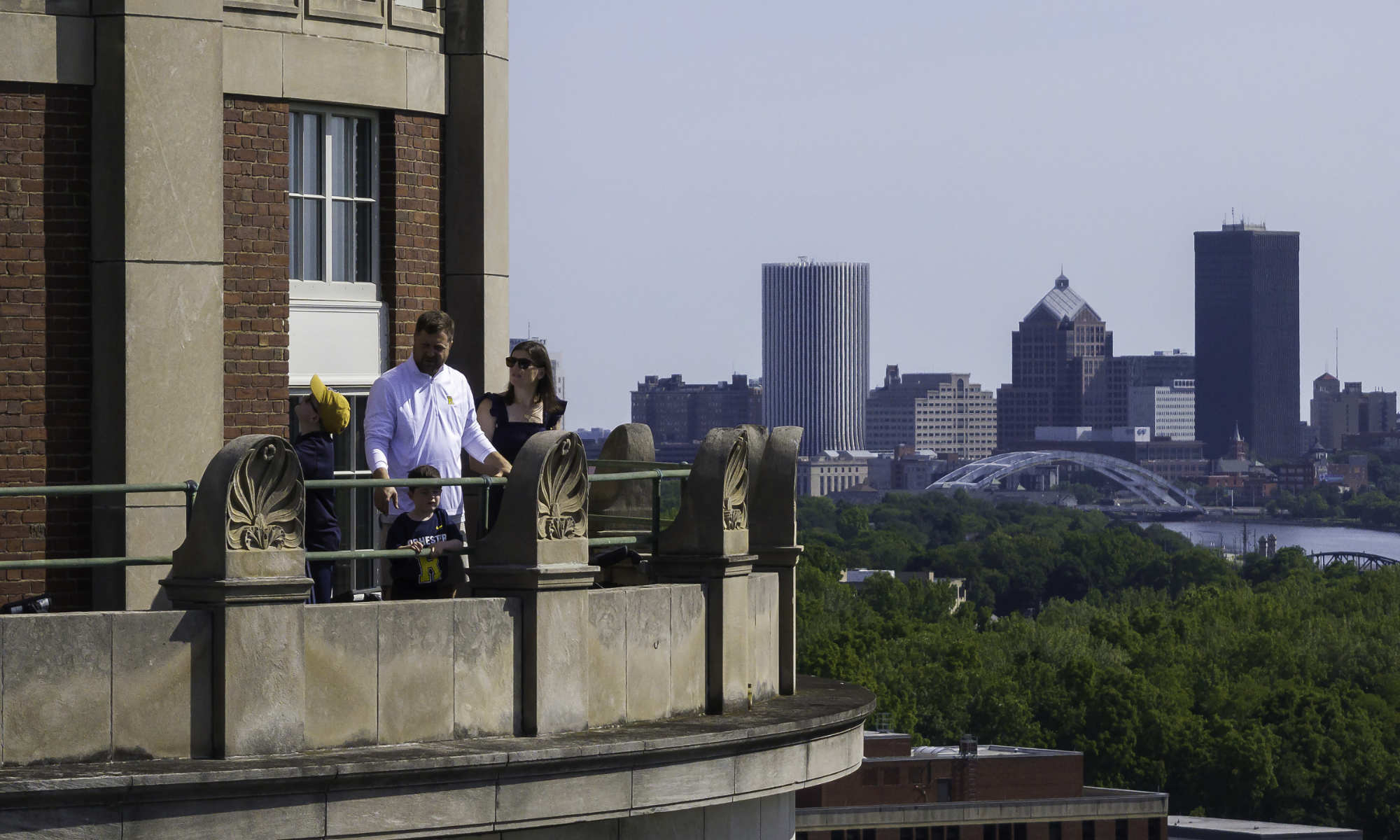 The Whites, a family of four, stand on the balcony of the Rush Rhees Library tower with the city of Rochester skyline in the background.