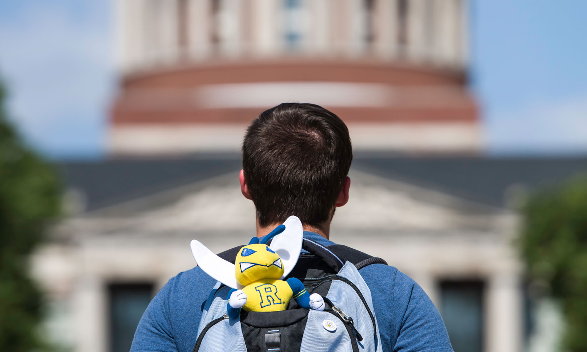 student facing Rush Rhees Library wears a backpack with a Rocky Yellowjacket mascot sticking out of it.