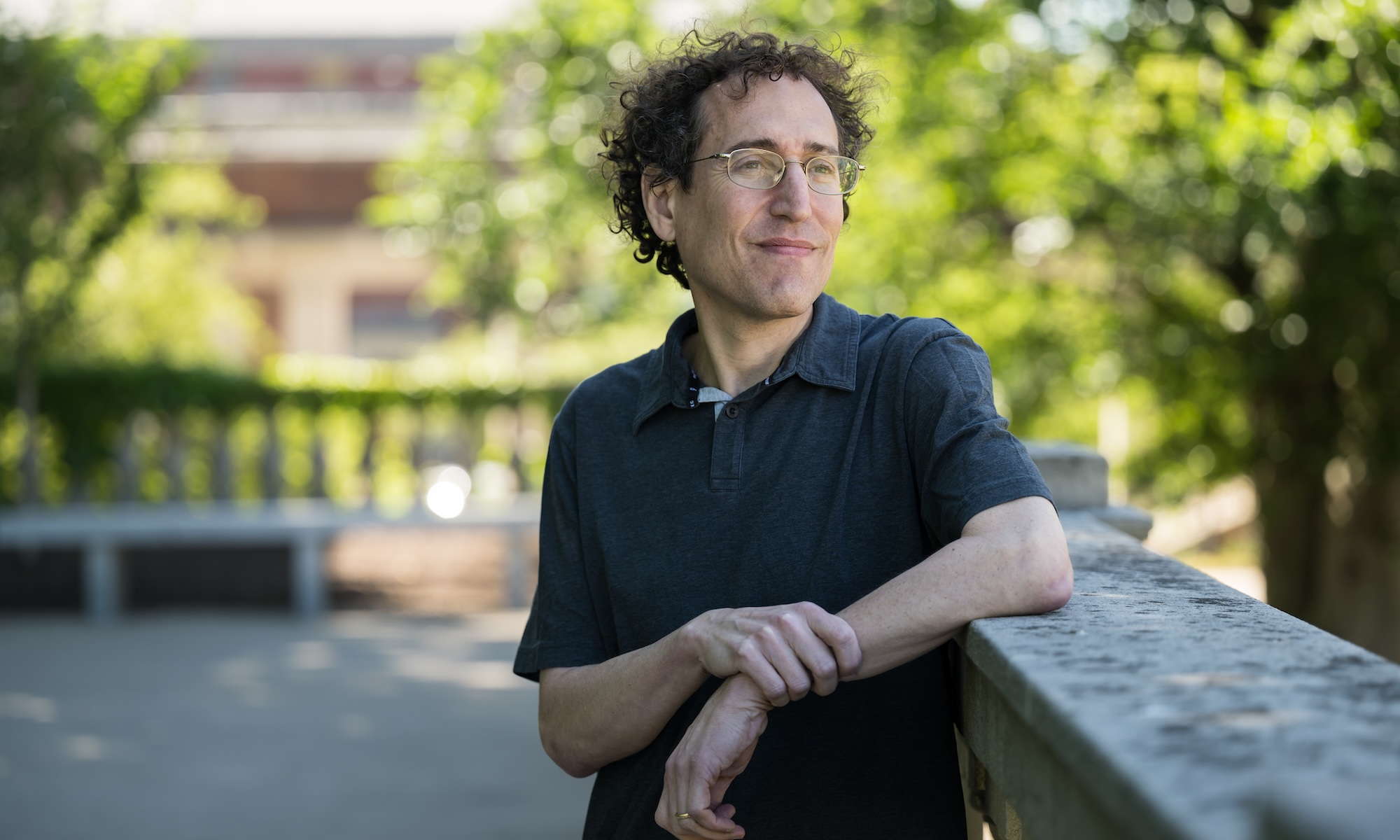 Druckman standing outdoors leaning against concrete wall with trees in background.