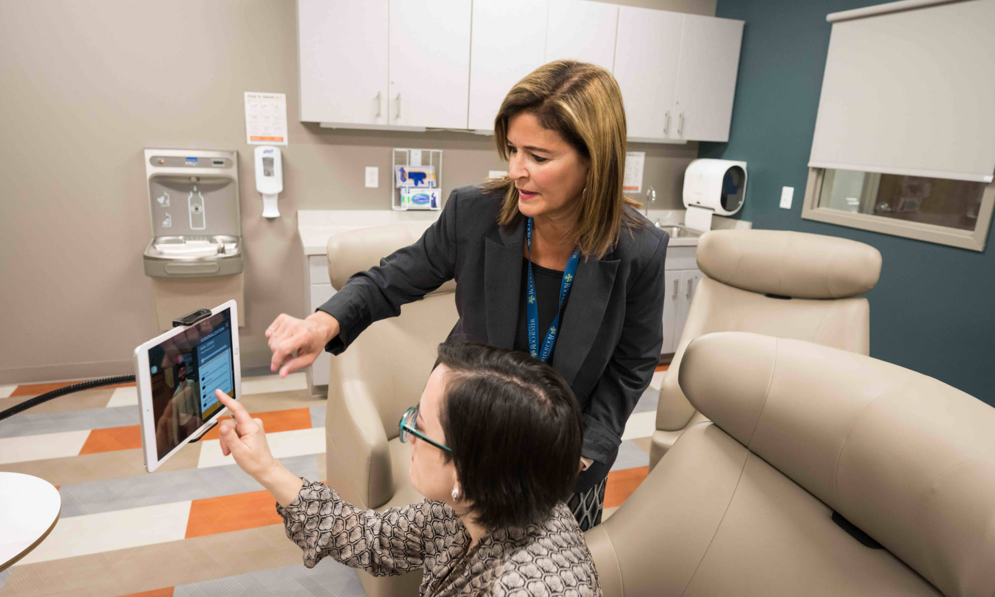 Two clinicians in a hospital-like setting touch the screen of a mounted tablet.