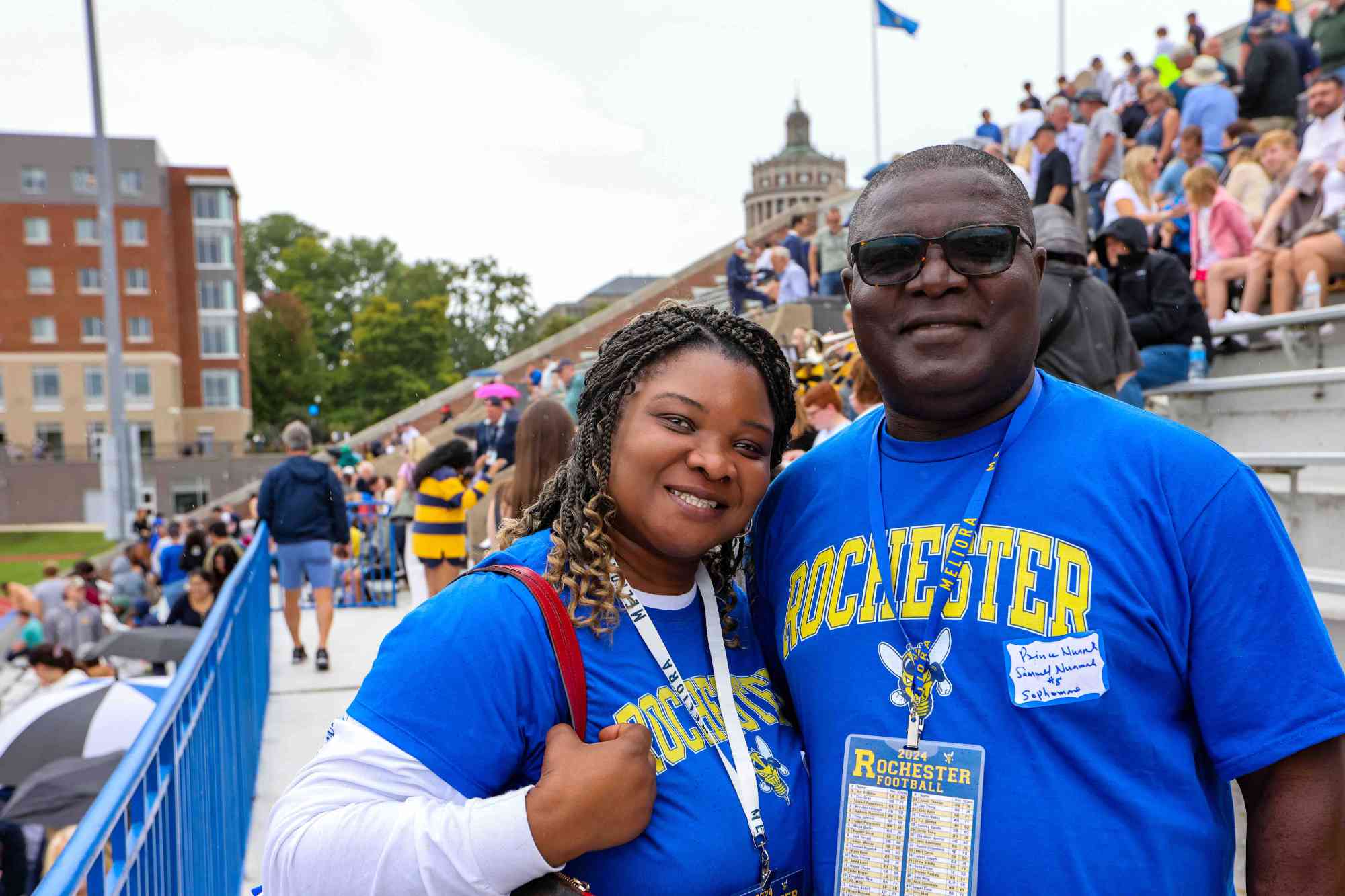 A father and daughter in matching University of Rochester t-shirts smile for a photo with bleachers and campus in the background.