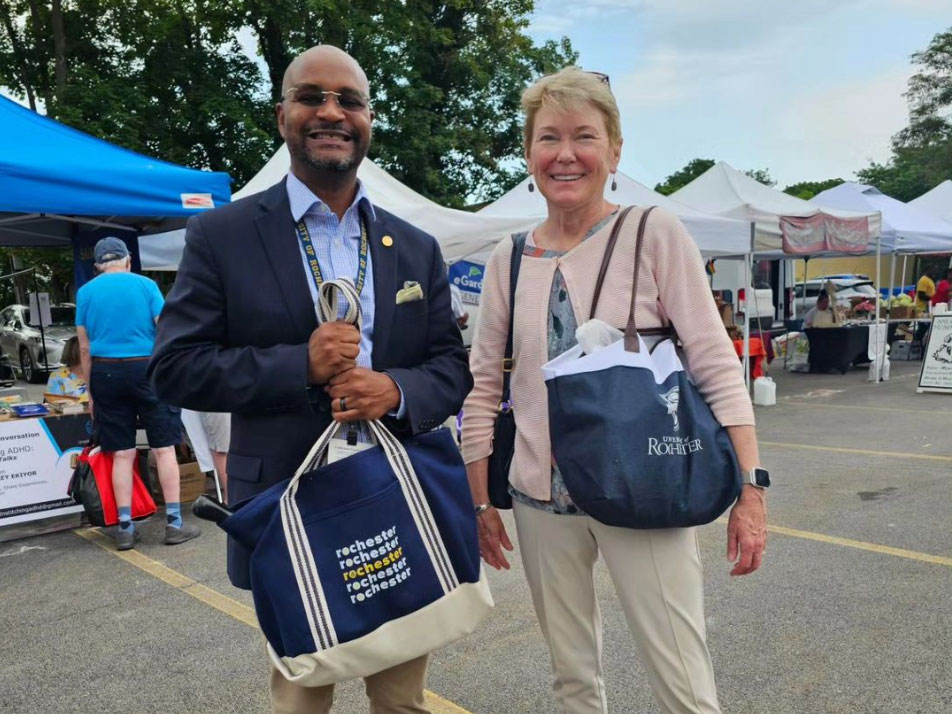 Two people smiling at a market with tents in the background, holding tote bags from the University of Rochester.