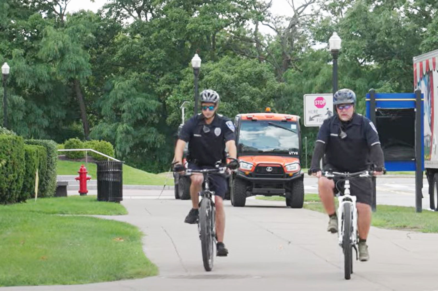 wo uniformed officers on bicycles ride towards the camera on a paved path, with an orange utility vehicle behind them and green trees in the background.