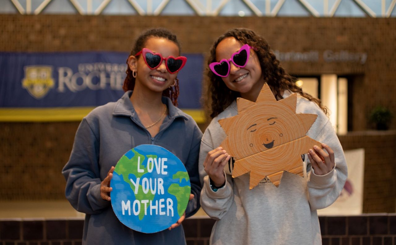 Two University of Rochester students wearing sunglasses indoors. One is holding a cardboard cutout of a sun, and the other is holding a sign of the Earth that says "Love your mother"