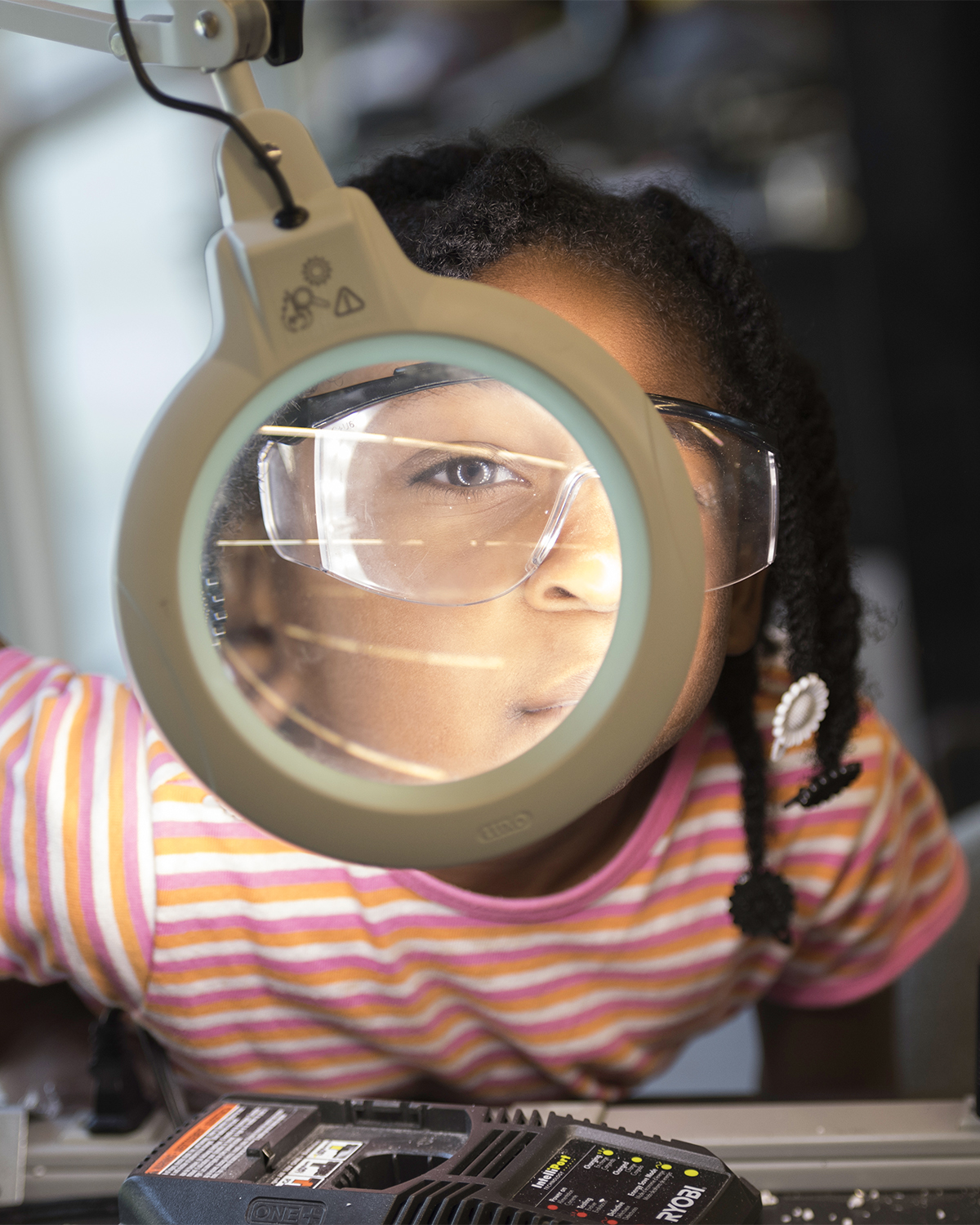 Young girl in safety glasses looking through an illuminated magnifying glass in the workshop in Rettner Hall.