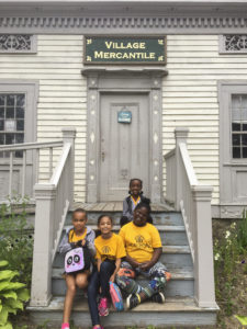 Students smiling on the steps of an old building with a sign on the building that reads "Village Mercantile" at Genesee Country Village and Museum.
