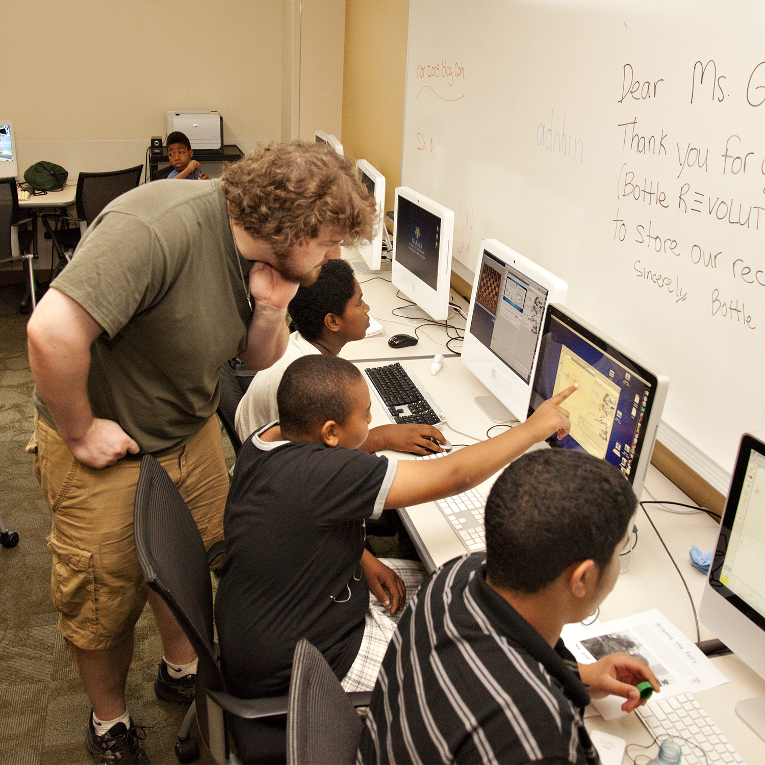 A teacher leans over to help a student working with others in the computer lab.