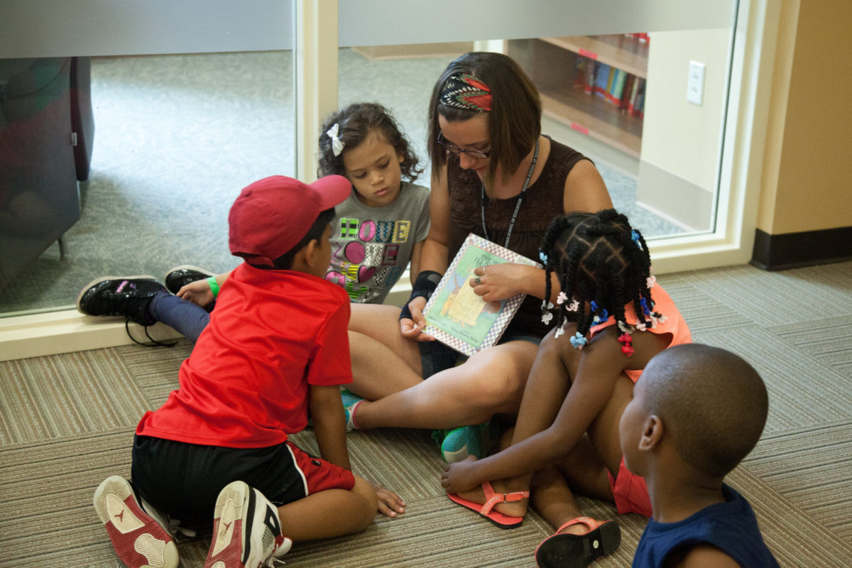 Woman sitting on the floor reading to a group of small children gathered closely around. her